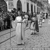 16. JUAN JOSÉ SERRANO GÓMEZ. Alegoría de la Virtud Teologal de la Fe en el cortejo de la Hermandad del Santo Entierro por la calle Alfonso XII. 1956.  La presencia de estas figuras simbólicas obedece al gusto romántico decimonónico de las hermandades. Tras la Fe, el grupo de los profetas, encabezado por el rey David con su arpa, añadido por primera vez en el Santo Entierro Grande de 1948. A la puerta de la iglesia de San Gregorio, los costaleros observan el desfile. © ICAS-SAHP, Fototeca Municipal de Sevilla, Archivo Serrano