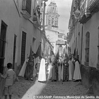 3. JUAN JOSÉ SERRANO GÓMEZ. Nazarenos de la Hermandad de la Hiniesta. 1930.  El tramo de nazarenos, con el estandarte de la hermandad conocido como “bacalao”, anuncia la llegada del paso de la Virgen de la Hiniesta por la estrechez de la calle Santa Paula. Al fondo, la espadaña del convento del mismo nombre. © ICAS-SAHP, Fototeca Municipal de Sevilla. Archivo Serrano