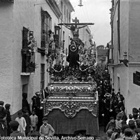 6. JUAN JOSÉ SERRANO GÓMEZ. Paso del Cristo de la Salud de la Hermandad de San Bernardo en la calle Gallinato. 1925-1927. El crucificado avanza por las estrechas calles del barrio de San Bernardo, uno de los arrabales históricos más populares. En este período le acompañaba la imagen de María Magdalena a los pies de la Cruz.   © ICAS-SAHP, Fototeca Municipal de Sevilla, Archivo Serrano